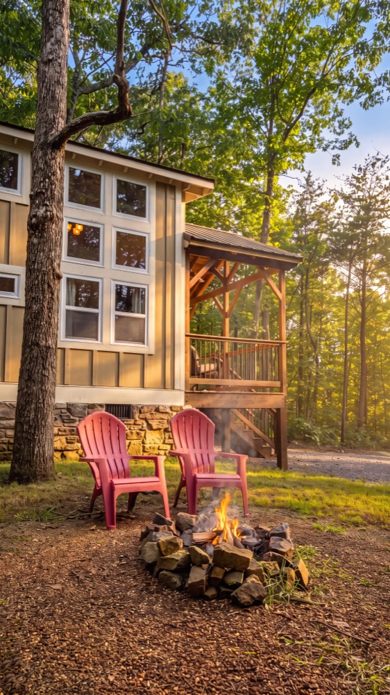 A stone fire ring with a small fire burning and two red Adirondack chairs beside it, in front of a board-and-batten cabin with a timber-frame covered stair landing.