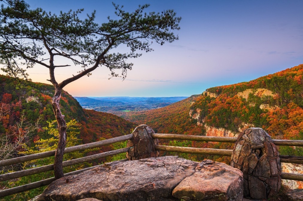 A rock overlook behind a split-rail fence at dusk, looking out across a canyon of red and orange autumn treetops.