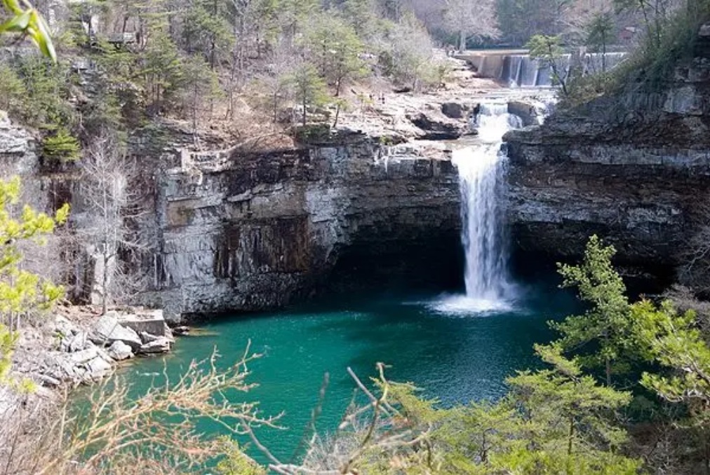 A tall waterfall dropping over a limestone bluff into a wide turquoise plunge pool framed by evergreens.