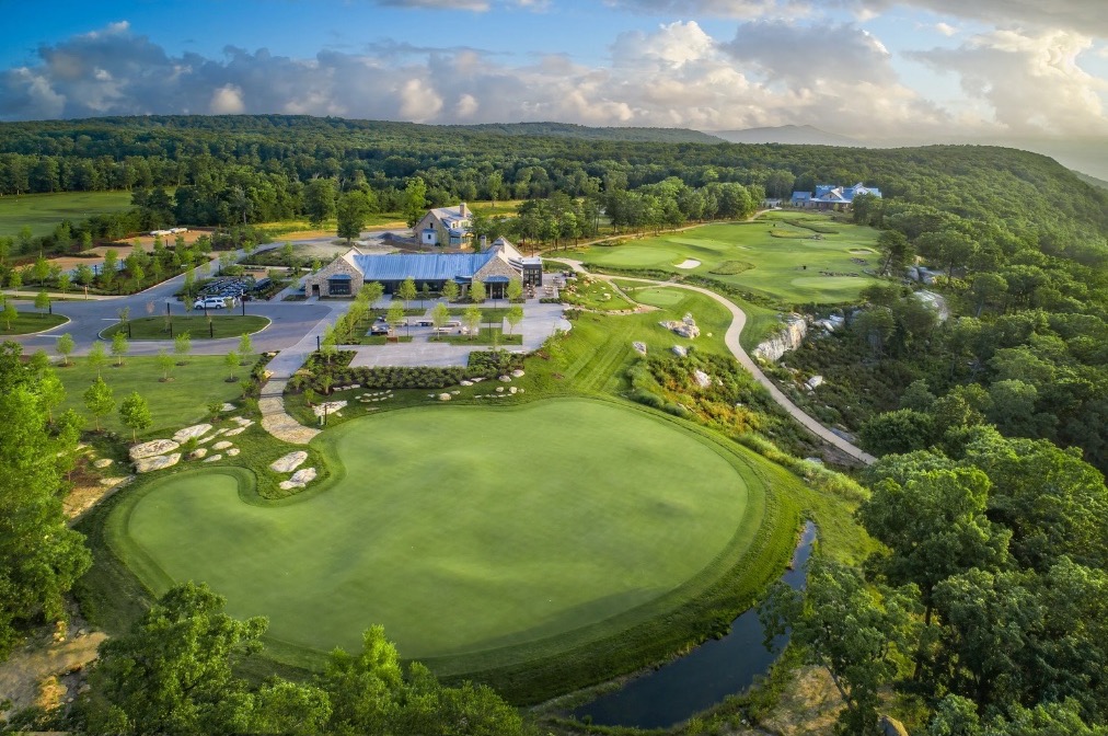 Aerial view of a mountaintop golf course, a broad manicured green ringed by woodland with the clubhouse and a long ridgeline beyond.