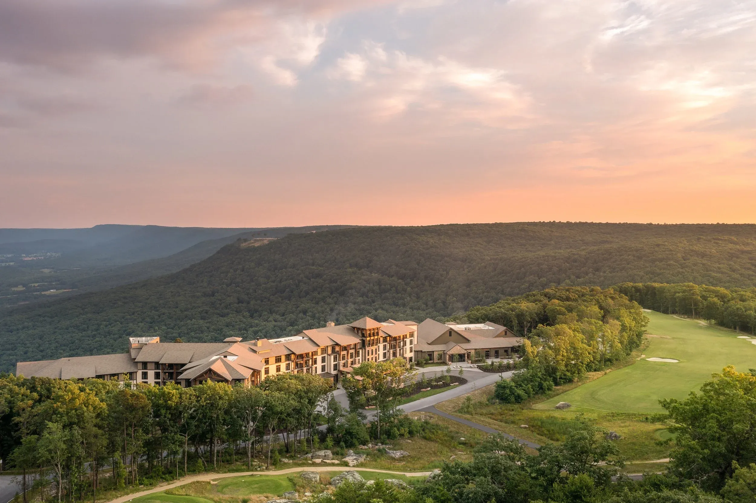 Aerial view of the McLemore Resort lodge on the brow of Lookout Mountain at sunset, a golf fairway beside it and forested ridgelines rolling away behind.