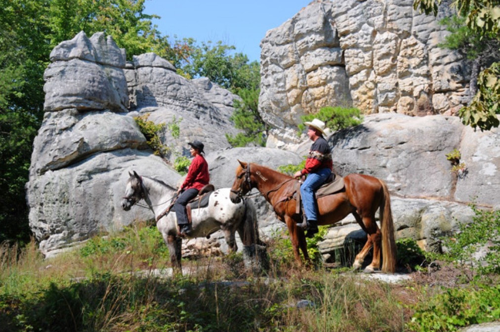 Two riders on horseback pausing beside weathered grey rock outcrops on a wooded trail.