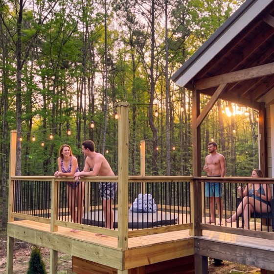 Guests talking on a cabin deck at sunset under string lights, a covered hot tub on the deck and woods beyond the rail.